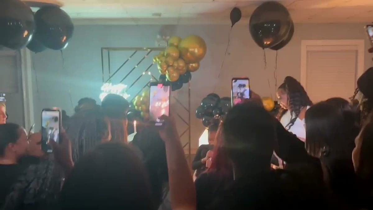 A private party room with black-and-gold balloons and a gold balloon arch, a crowd holding up phones to film