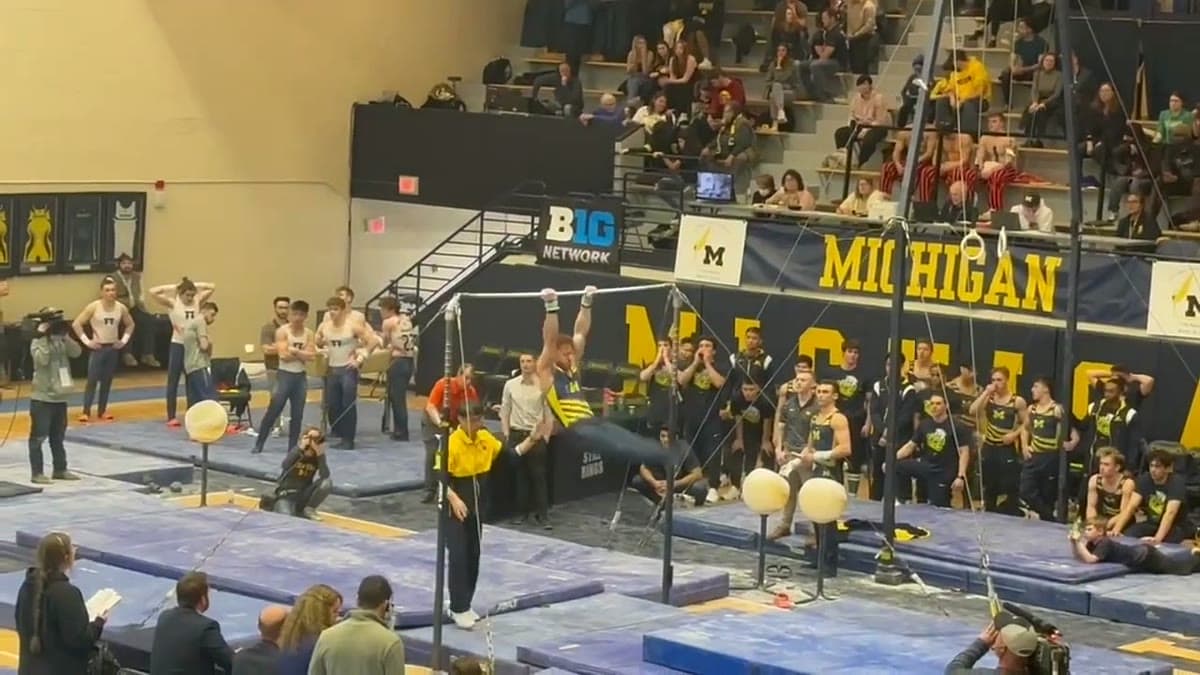 A Michigan gymnast competing on the high bar at Cliff Keen Arena while the team watches from the floor