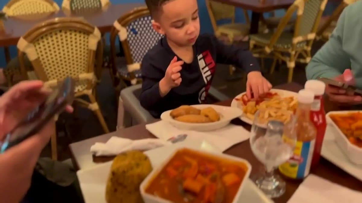 Plates of jollof rice, stewed meat and fried sides on a table at a Dominican restaurant, with a child eating in the background