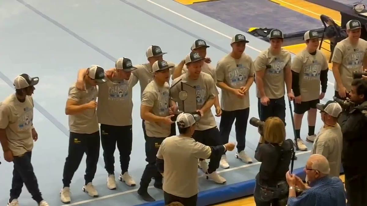 The current Michigan men's gymnastics team celebrating with a trophy on the competition floor